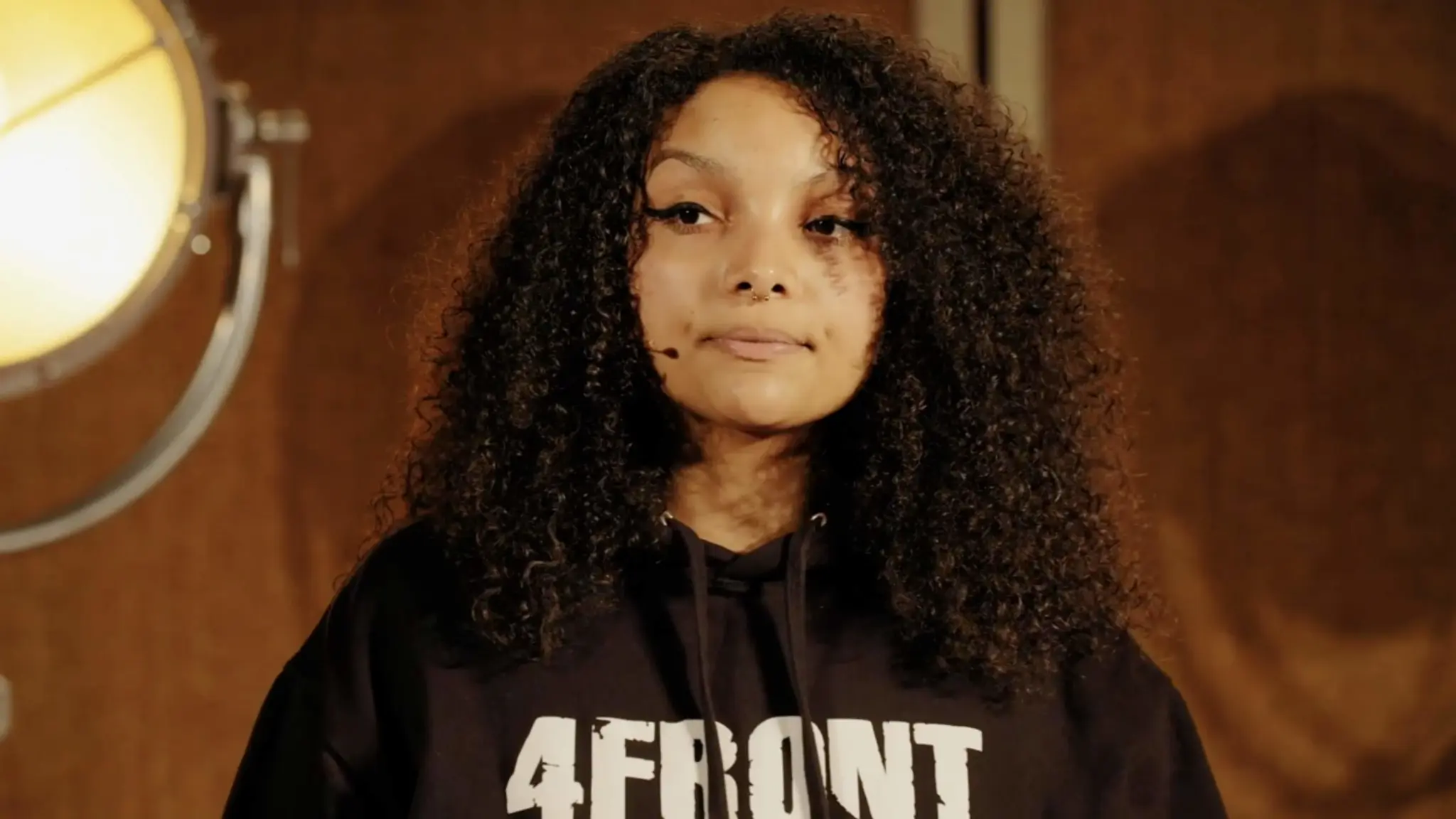 A screenshot from the video 'Why fighting for racial equity means reimagining justice' from Youtube. The image is of Temi Mwale the speaker who is a black woman with curly hair down to her shoulders. She is wearing a black hooded jumper with a '4Front' logo. She is standing on a stage with a stage light behind her on the left.