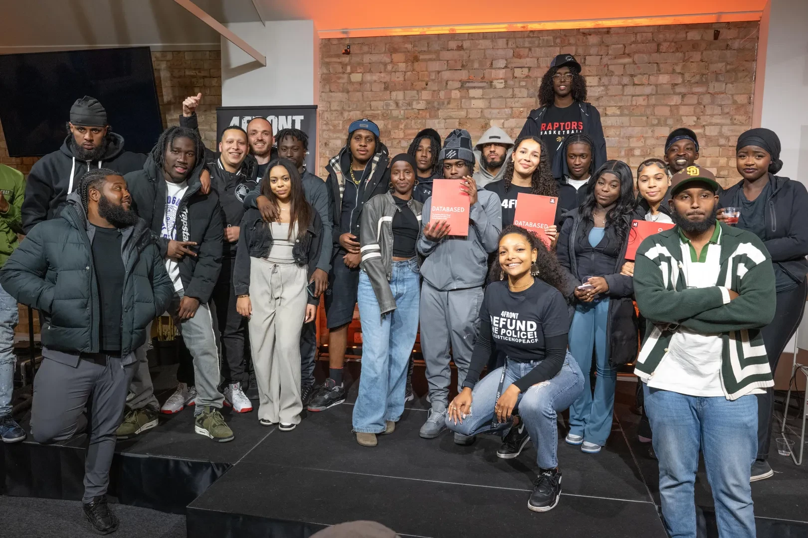 A large group of young black and brown people are standing in front of a brick wall in an interior. A few of them are holding a red booklet titled "DATABASED - The Impacts of Racist 'Gang' Profiling and Surveillance on Young People in London".