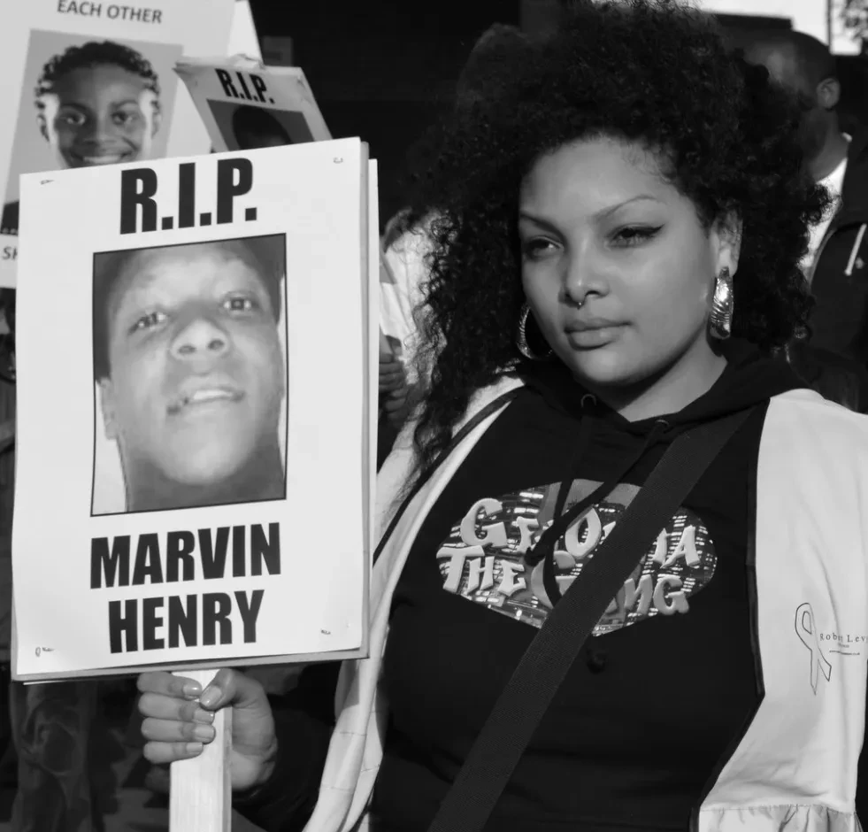 A black and white shot of a young person at a protest, holding a sign that reads "R.I.P. Marvin Henry" with a photo of the victim. Other signs are also discernible in the background.