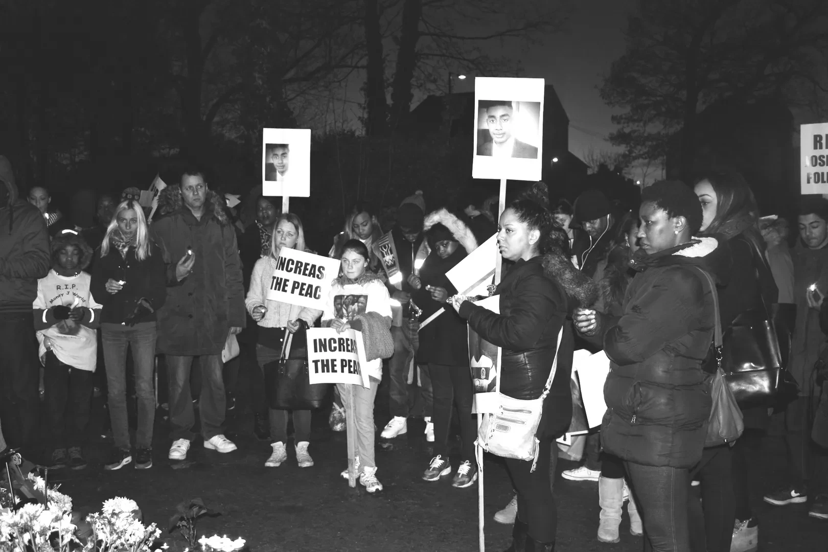 Black and white shot of a large peaceful protest with many people standing in silence with a solemn expression. Some of them are holding signs that read "Increase the peace" and others photographs of victims.
