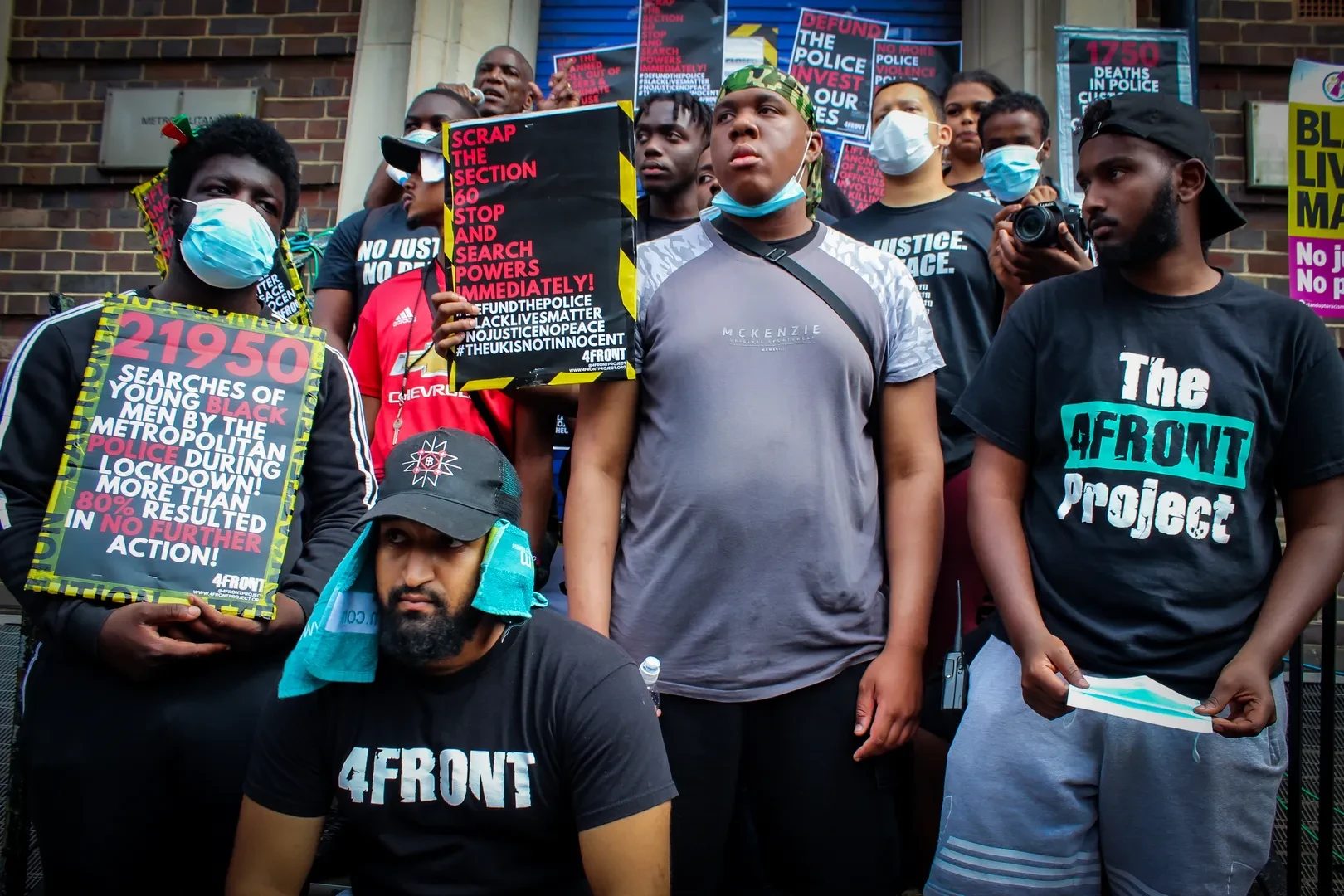 A group of people of colour, many of whom are wearing face masks, standing in front of a brick wall, holding many signs that denounce racisme and police brutality during lockdown.