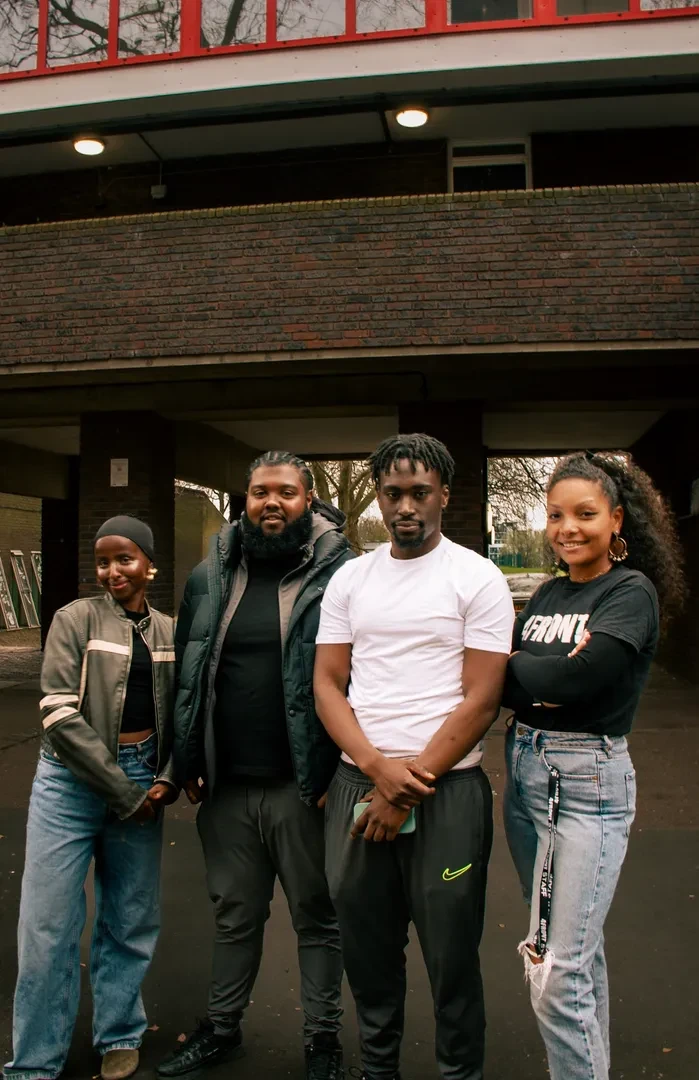 Small group of four young people standing in an urban environment, facing the camera with a smile.