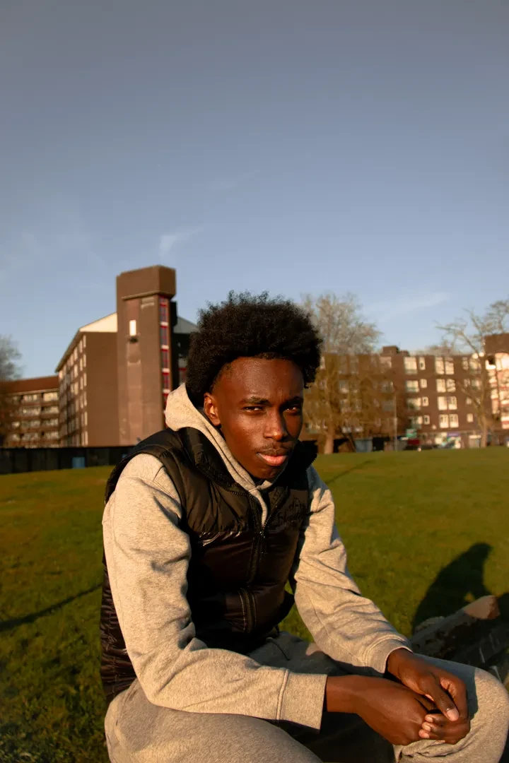 Young person sitting on the grass in a park, facing the camera, with brick buildings further in the background.