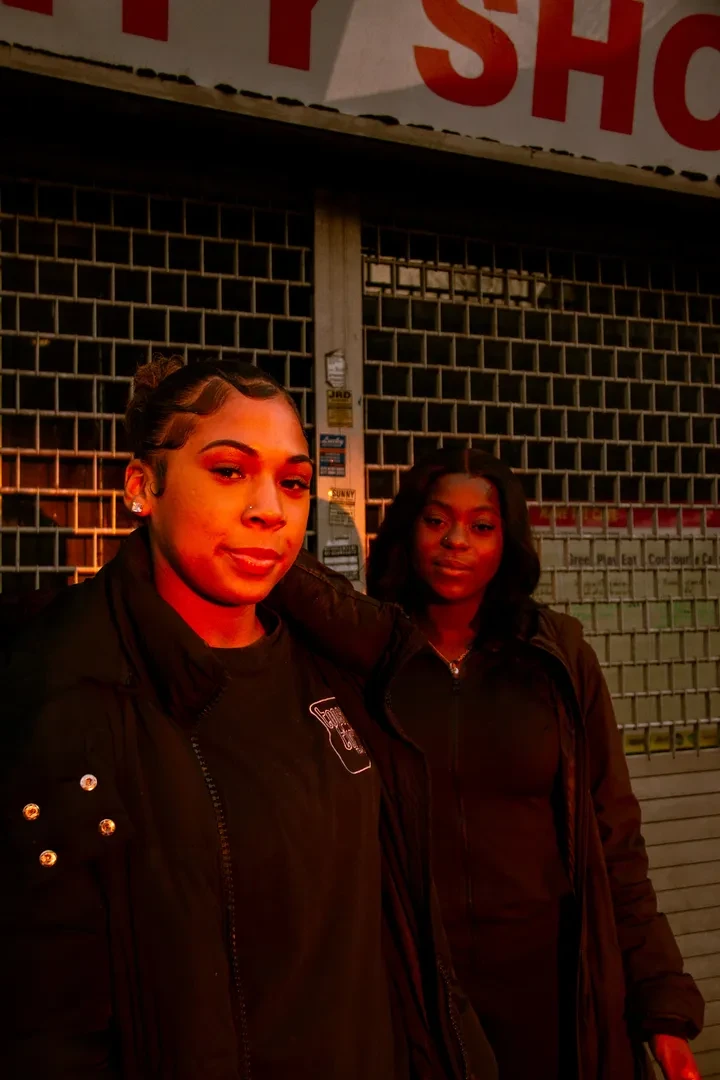 Two young people lit with evening light, standing in front of a building with a neutral expression.