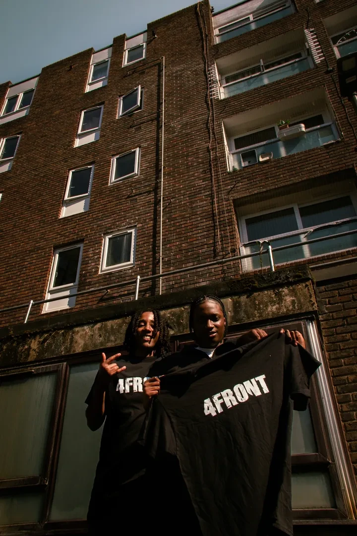 Two young people standing in front of a brick building. The one on the front is holding a black t-shirt with "4FRONT" written on it with white character, and the one behind is wearing that same t-shirt.