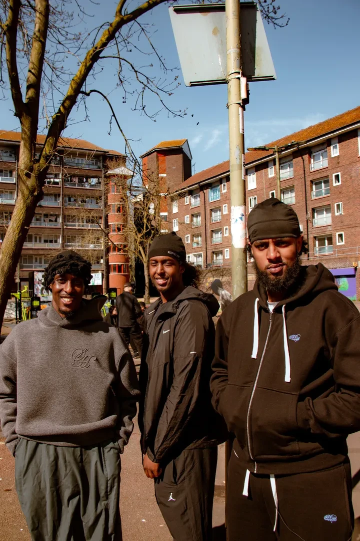Three young people standing in an urban environment, looking at the camera with a smile.