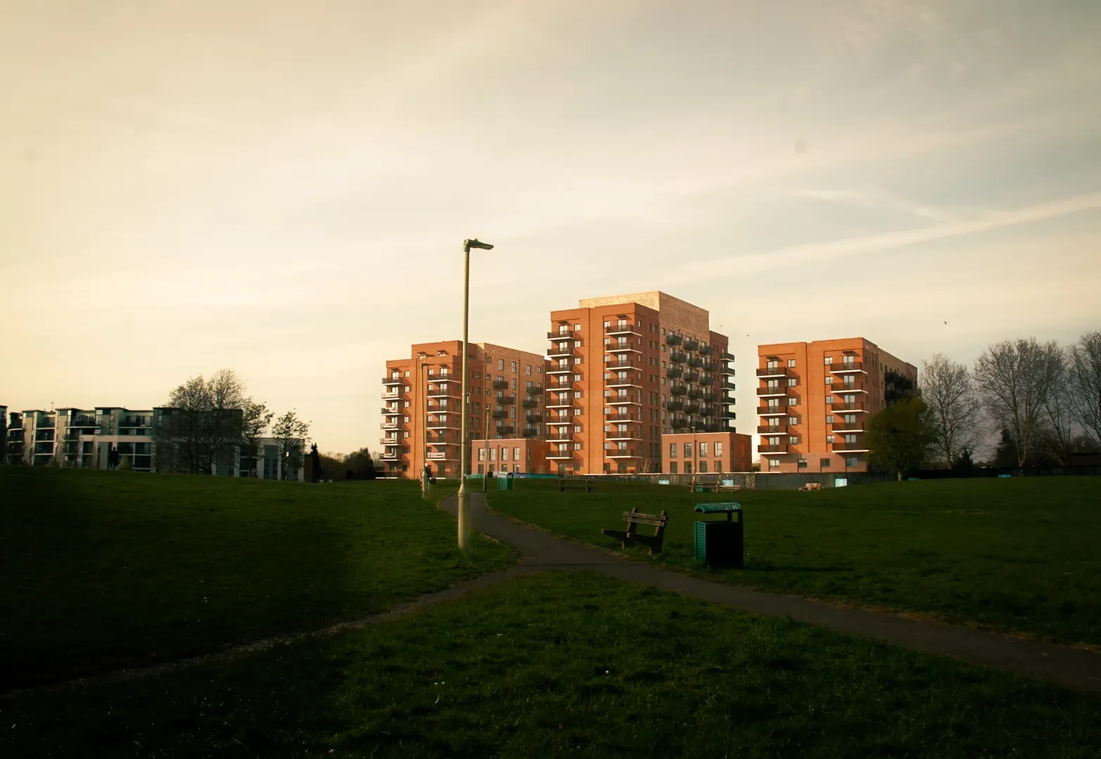 Large environment shot of a grassy park and several recent buildings forming a gentrified neighborhood in the background.