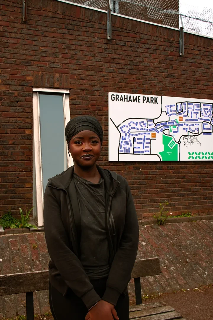 A young person dressed in black is standing in front the map sign for Grahame Park on a brick building, facing the camera