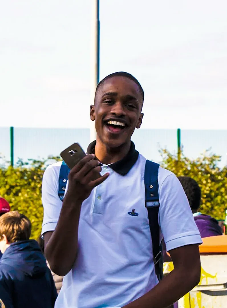 A laughing young person in a white polo wearing a backback, holding a smartphone in their right hand and facing the camera.