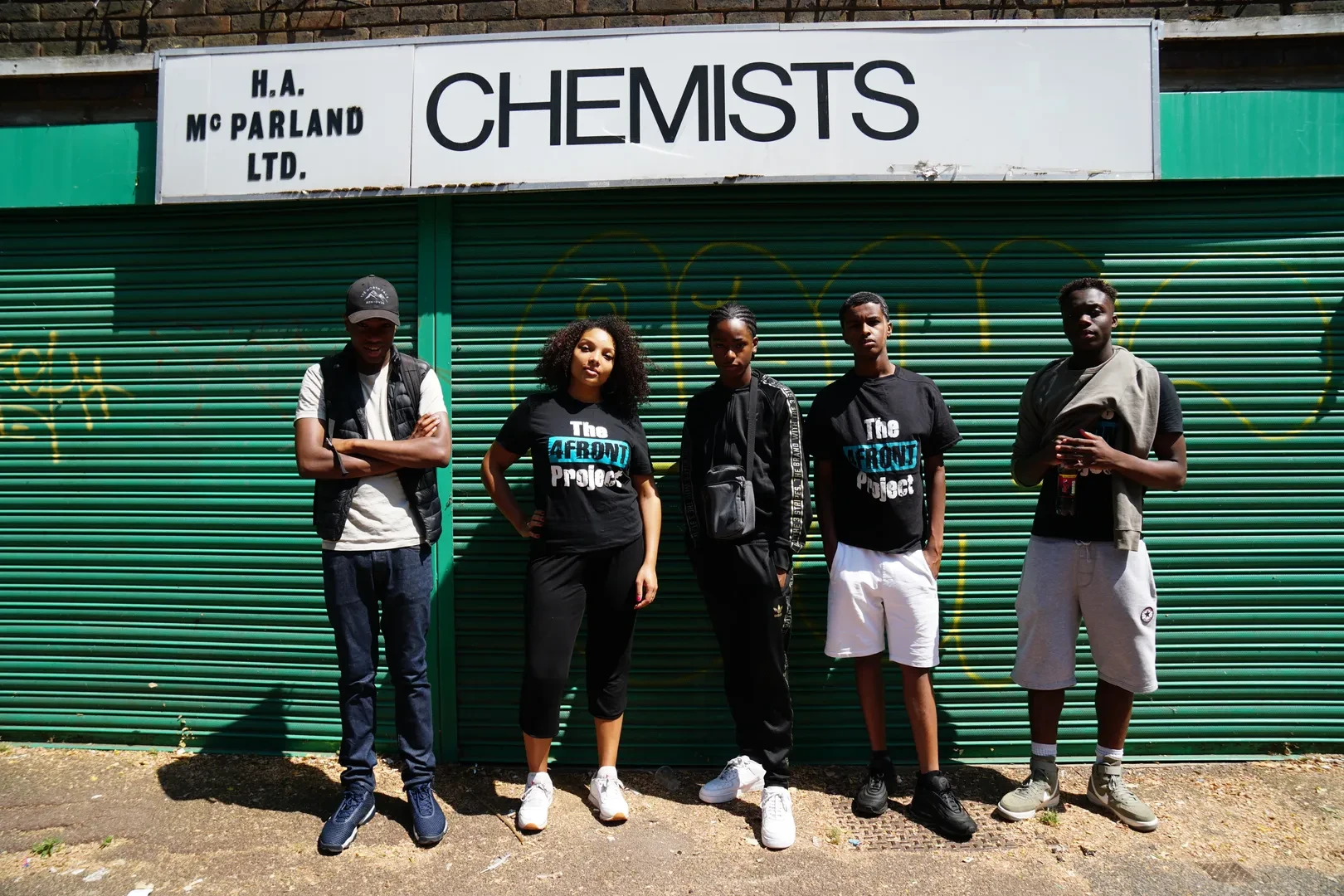 A small group of five young people of color, standing in front of a closed green shutter with graffiti and a "CHEMISTS" sign above them. Two of them are wearing a black t-shirt with a sign that reads "The 4FRONT Project".