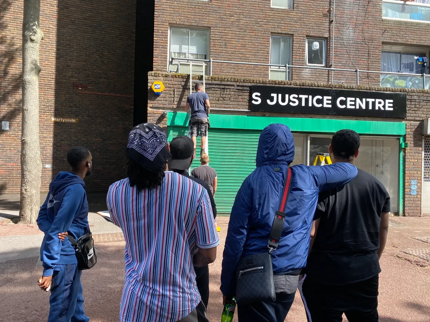 A small group of five people, turning their back to the camera, looking at Jahiem's Justice Centre while a worker on a ladder is working on the Centre's sign.