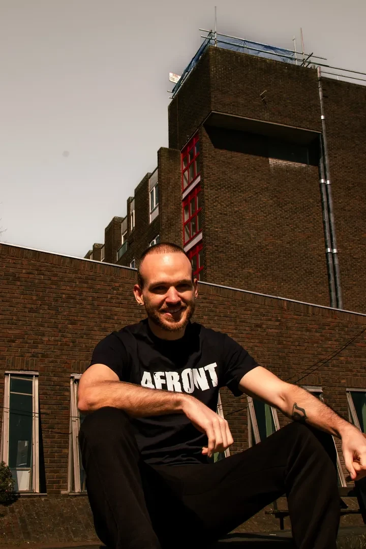 A young person is sitting on the ground in front of a brick building, looking at the camera with a smile and wearing a black t-shirt with "4FRONT" written on it with white characters.