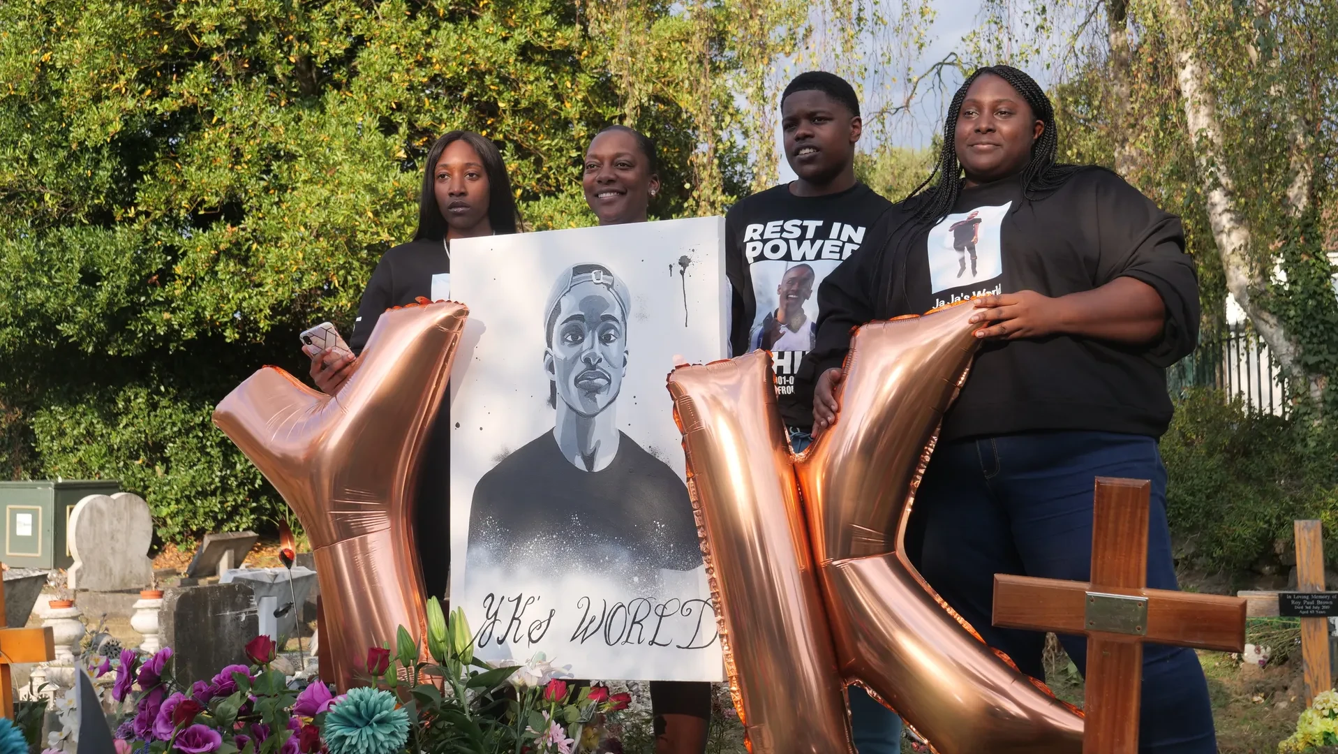 Small group of 4 young people in a graveyard, holding a large black and white portrait painting of their deceased young relative with "YK's World" written underneath. They are also holding two large helium balloons shaped like the letters Y and K.