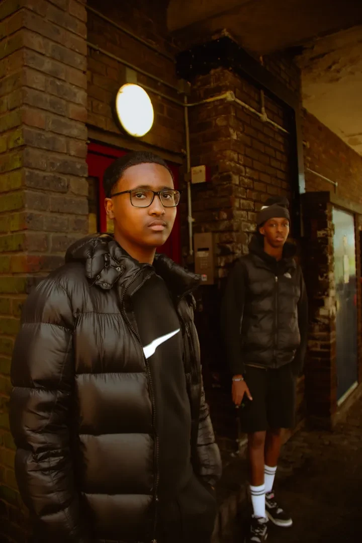 Two young people in urban attire stand in a dimly lit alley, looking at the camera with a neutral expression.