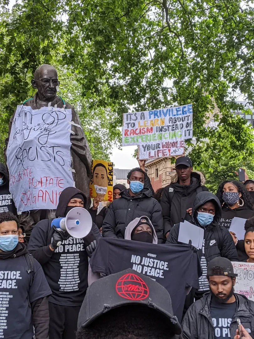Large group of people in black hoodies during a BLM protest, holding and wearing t-shirts that read "No Justice, No Peace" and wearing face masks. They stand in front of a statue of Gandhi, on which is stuck a sign that reads "Stop glorifying Racists - Educate, Agitate, Organise"