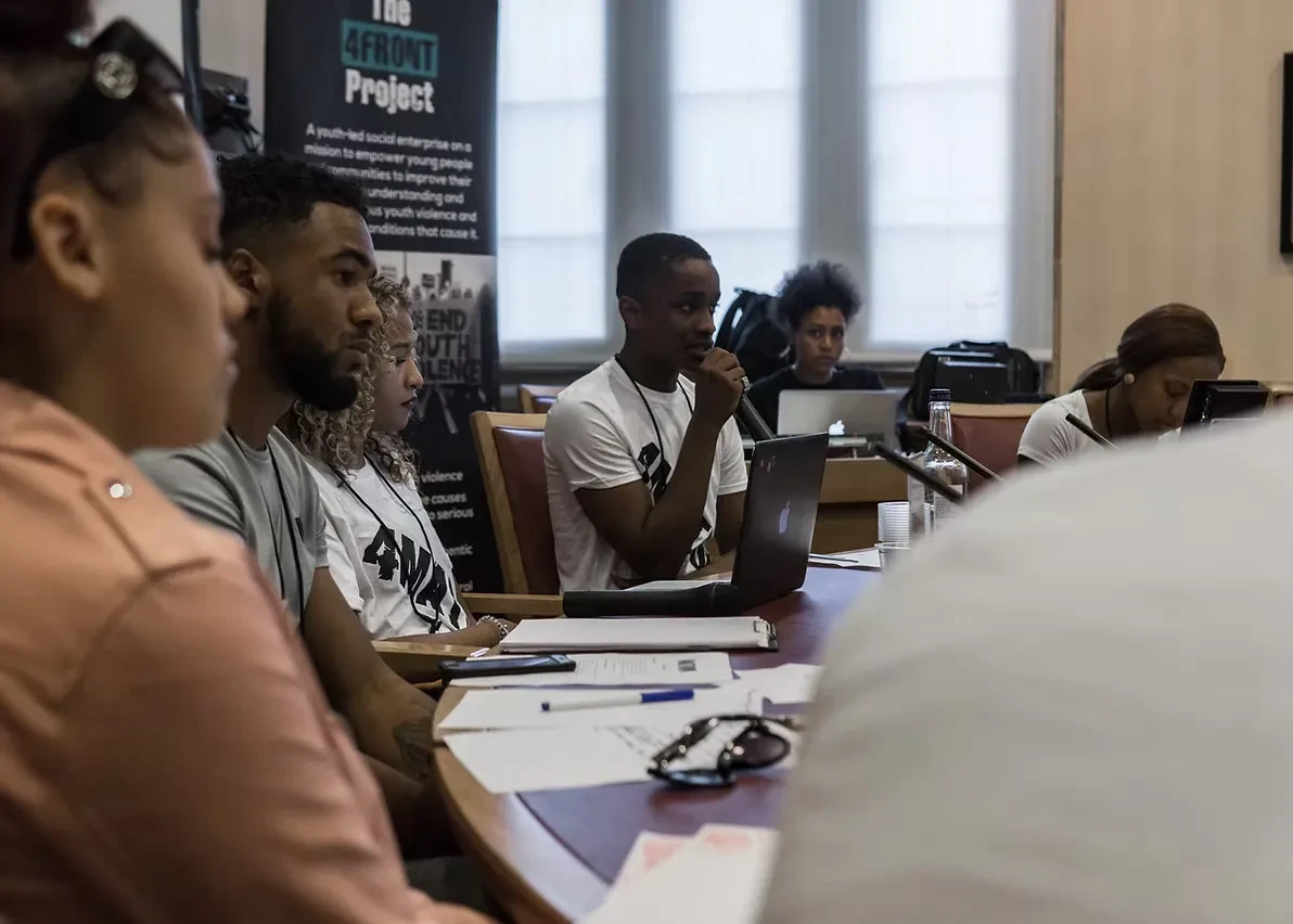 A group of young people sit around a conference table with papers, laptops and microphones, with a 4Front banner standee behind them.