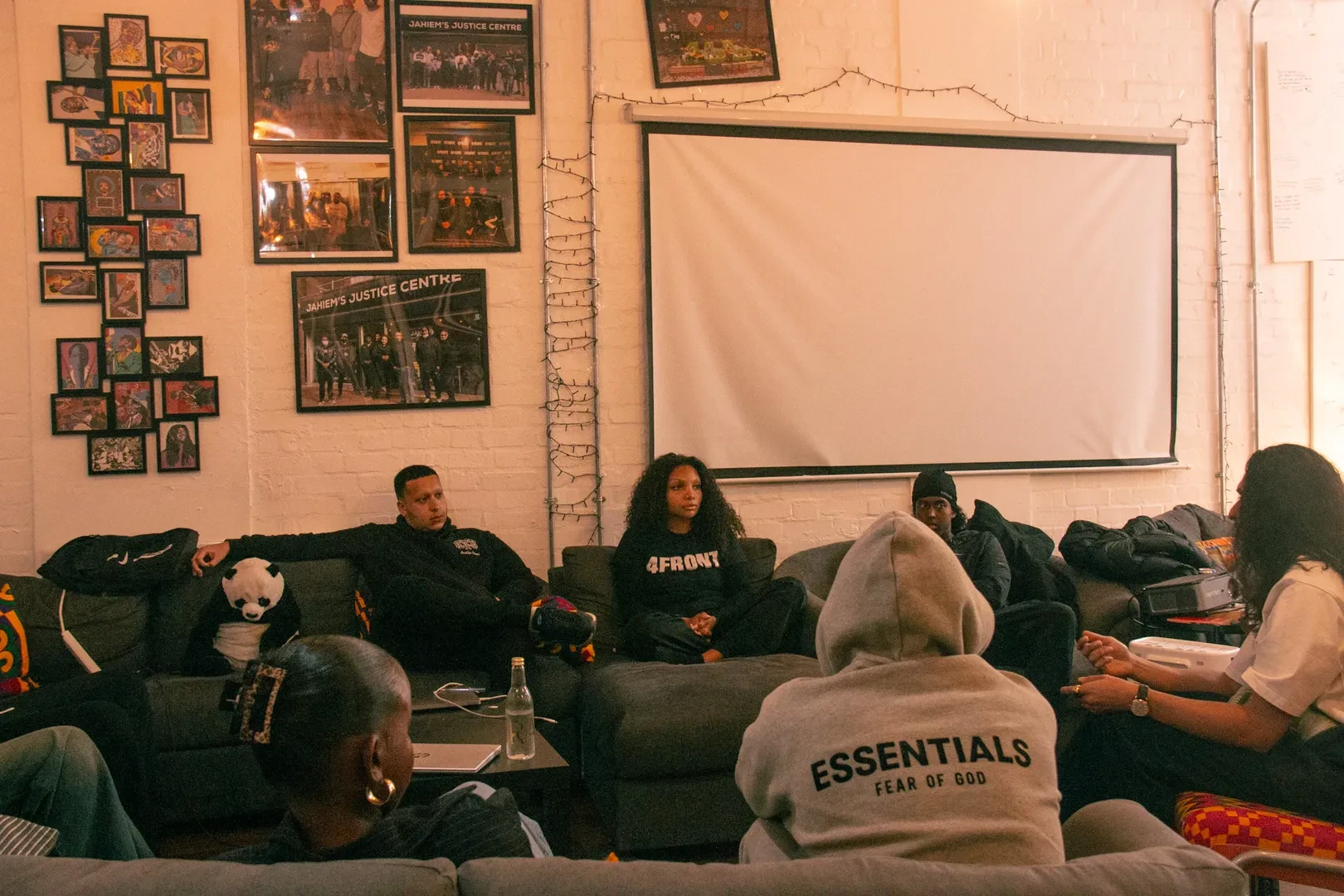 Small group of young people gathered in a dimly lit break room in Jahiem's Justice Centre, sitting on sofas around coffee tables, in front of a white wall decorated with various framed photographs and one large projector screen.