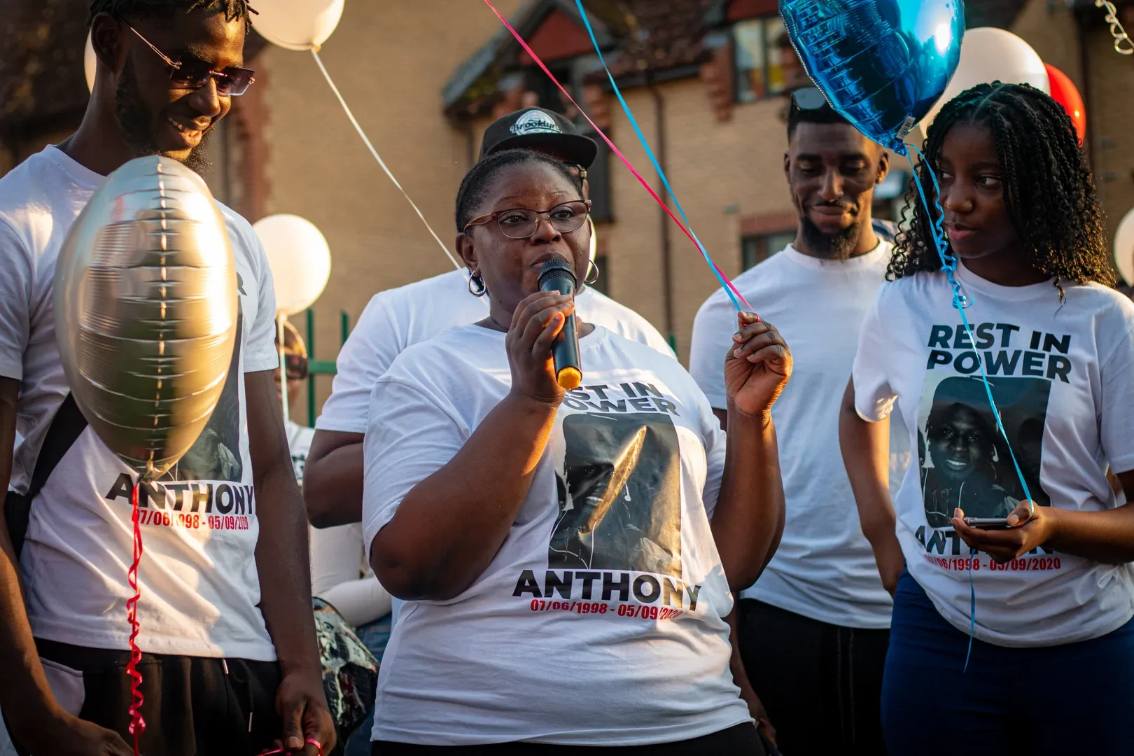 A group of people holding party helium balloons and wearing white t-shirts that read "Rest in Power Anthony - 07/06/1998 - 05/09/2020" with a portrait photo of the victim. The person in the middle is speaking in a microphone.
