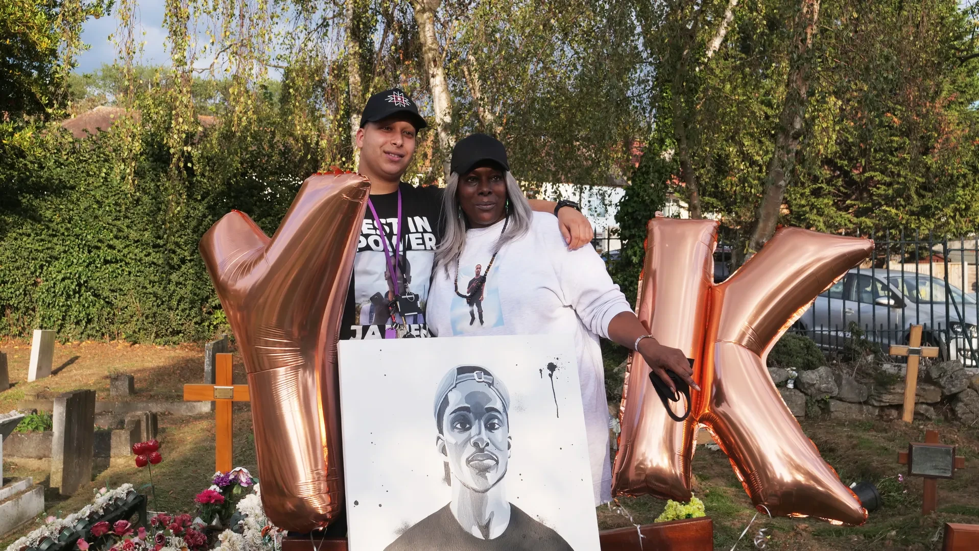 Two young people are standing in a graveyard, surrounded by two large helium helium balloons shaped like the letters Y and K. They are holding a large black and white portrait painting of a young person.
