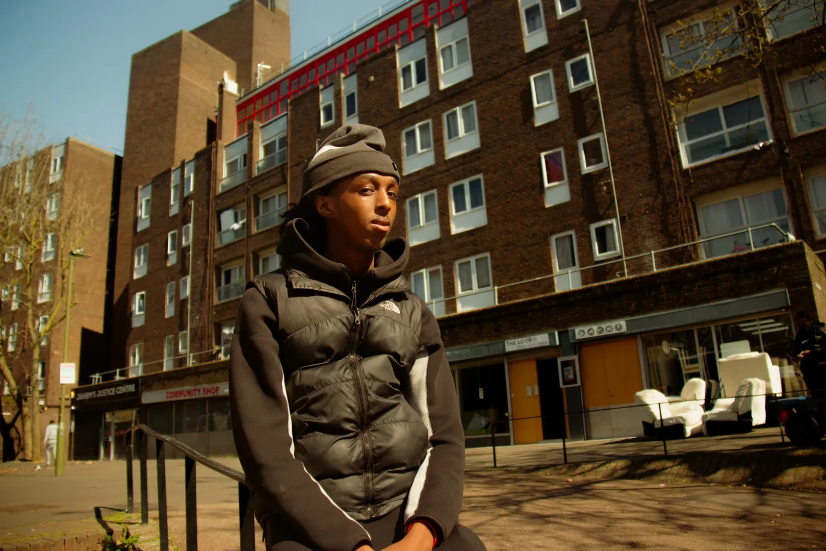 A young person is standing in front of a building block, looking at the camera with a neutral expression.