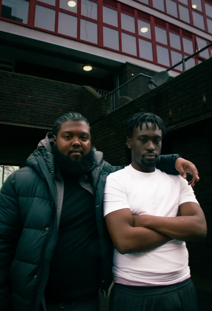 Two young people are standing in a dimly lit urban environment with brick walls.They are looking at the camera with a neutral expression.