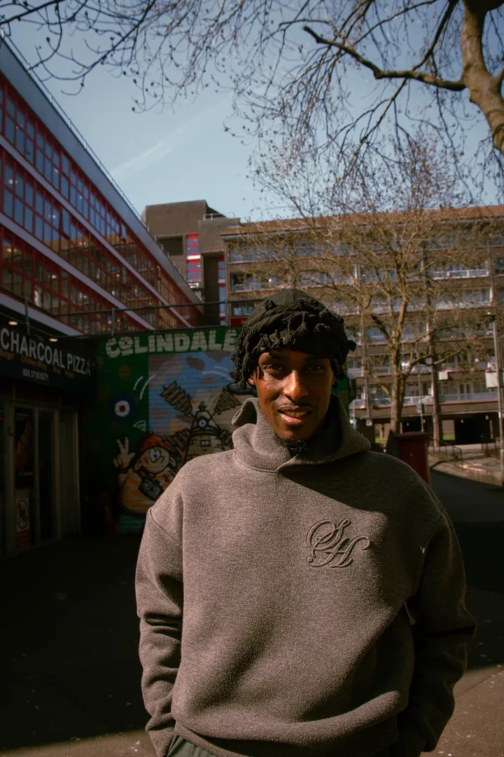 A young person is standing in an urban environment, looking at the camera smiling.