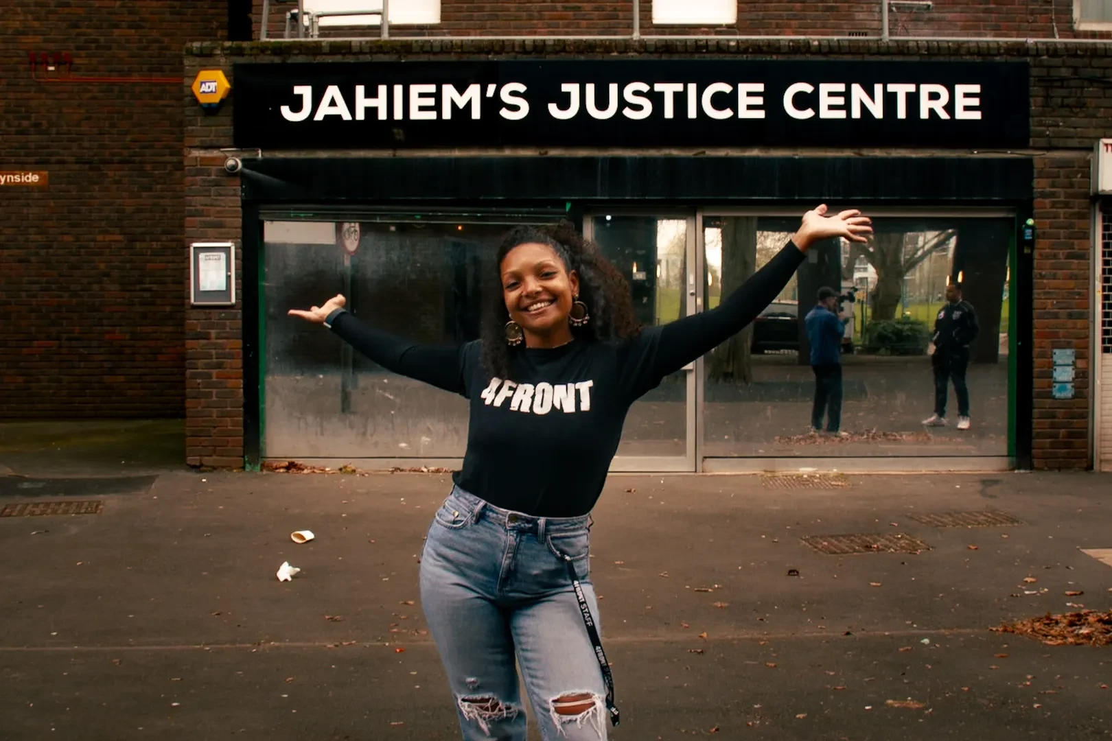 A smiling young person with a black "4Front" shirt and arms outstretched is standing in front of Jahiem's Justice Centre.
