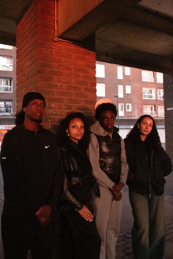 Four young people with a solemn expression are standing close to each other in a dimly lit alleyway.
