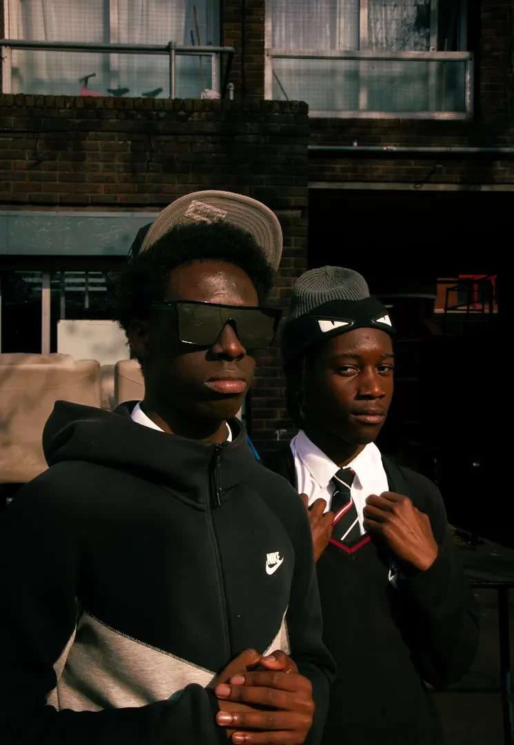 Two young people are standing in front of a building block with a serious expression. One of them is wearing sunglasses, and the other one a cravat tie.