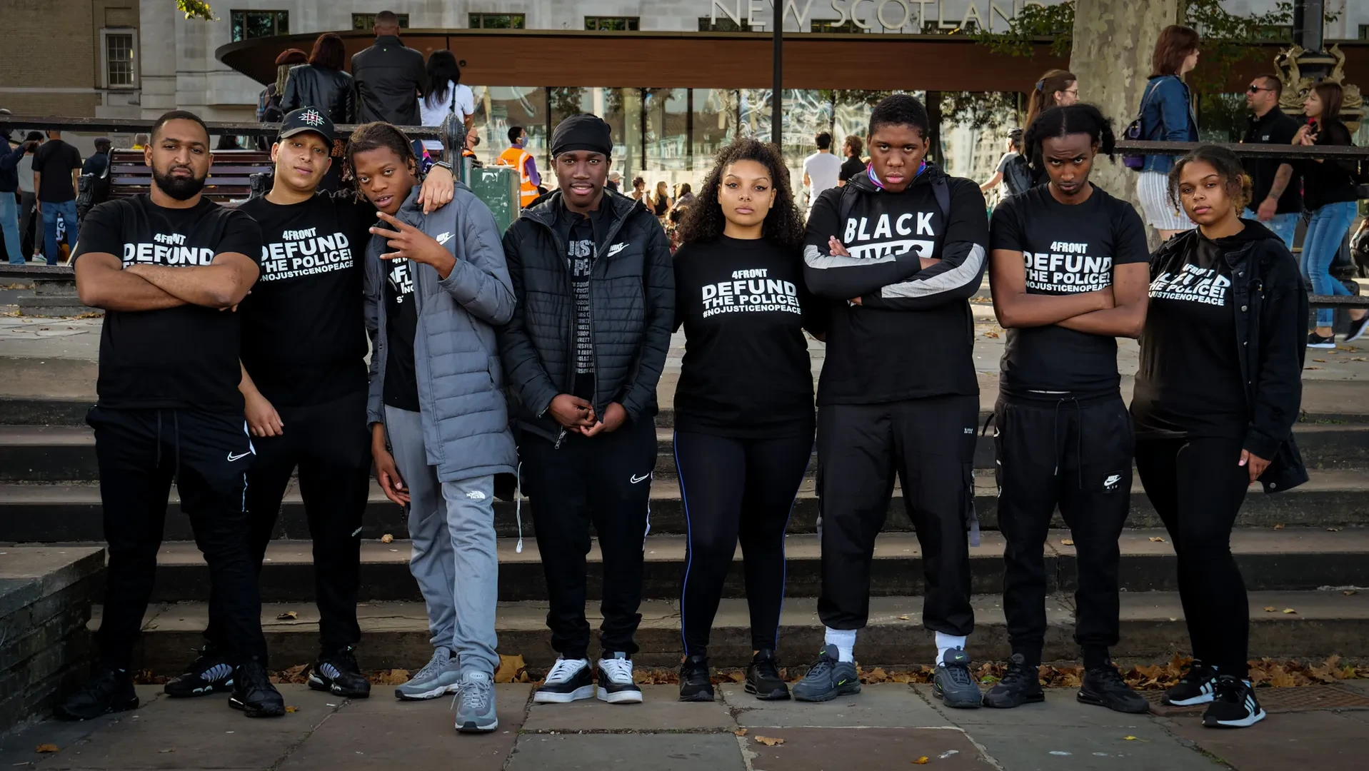 A group of eight young people are standing next to each other with a serious expression. They are wearing black t-shirts that bear inscriptions like "Defund the Police" and "Black Lives Matter".